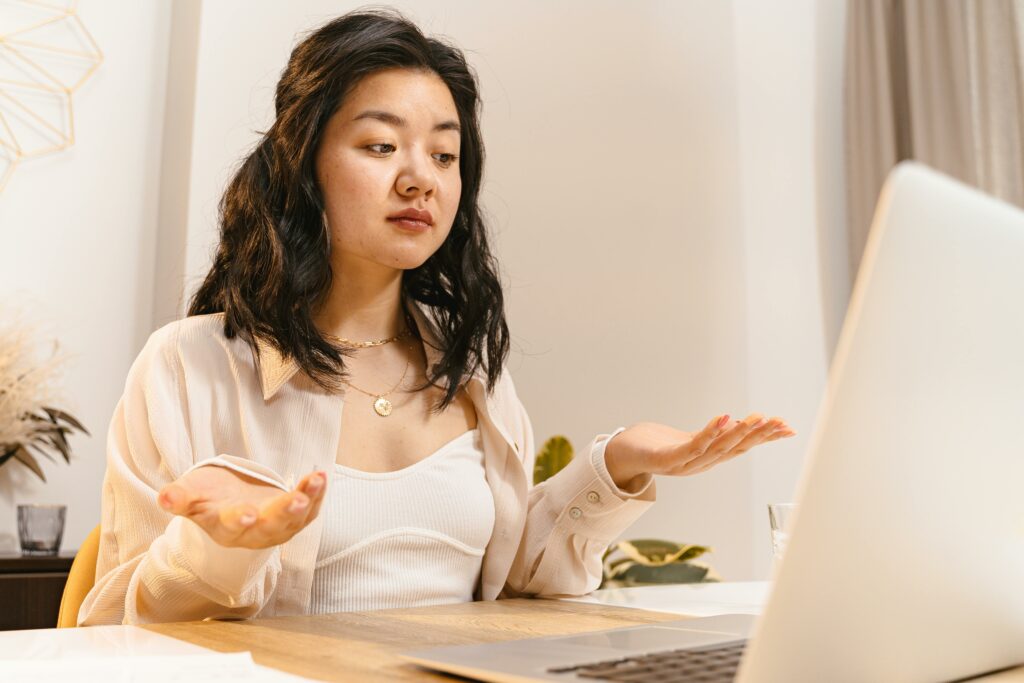 Asian woman in a long sleeve shirt, appearing puzzled while sitting indoors and looking at a laptop.